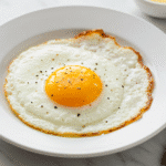 Perfect over easy egg with golden runny yolk on white plate, seasoned with black pepper and salt